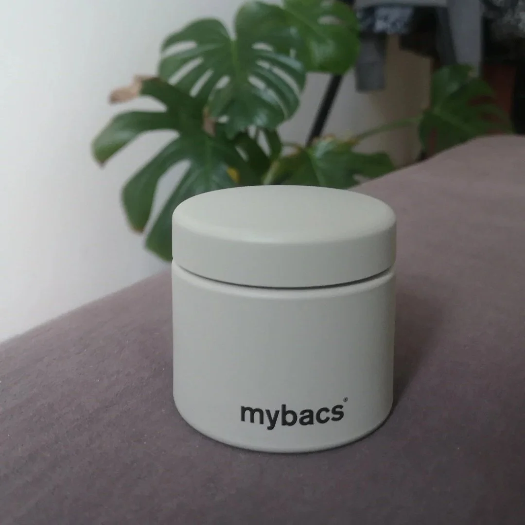 A white cylindrical container with a lid, labeled 'mybacs', placed on a gray surface with a green houseplant in the background.
