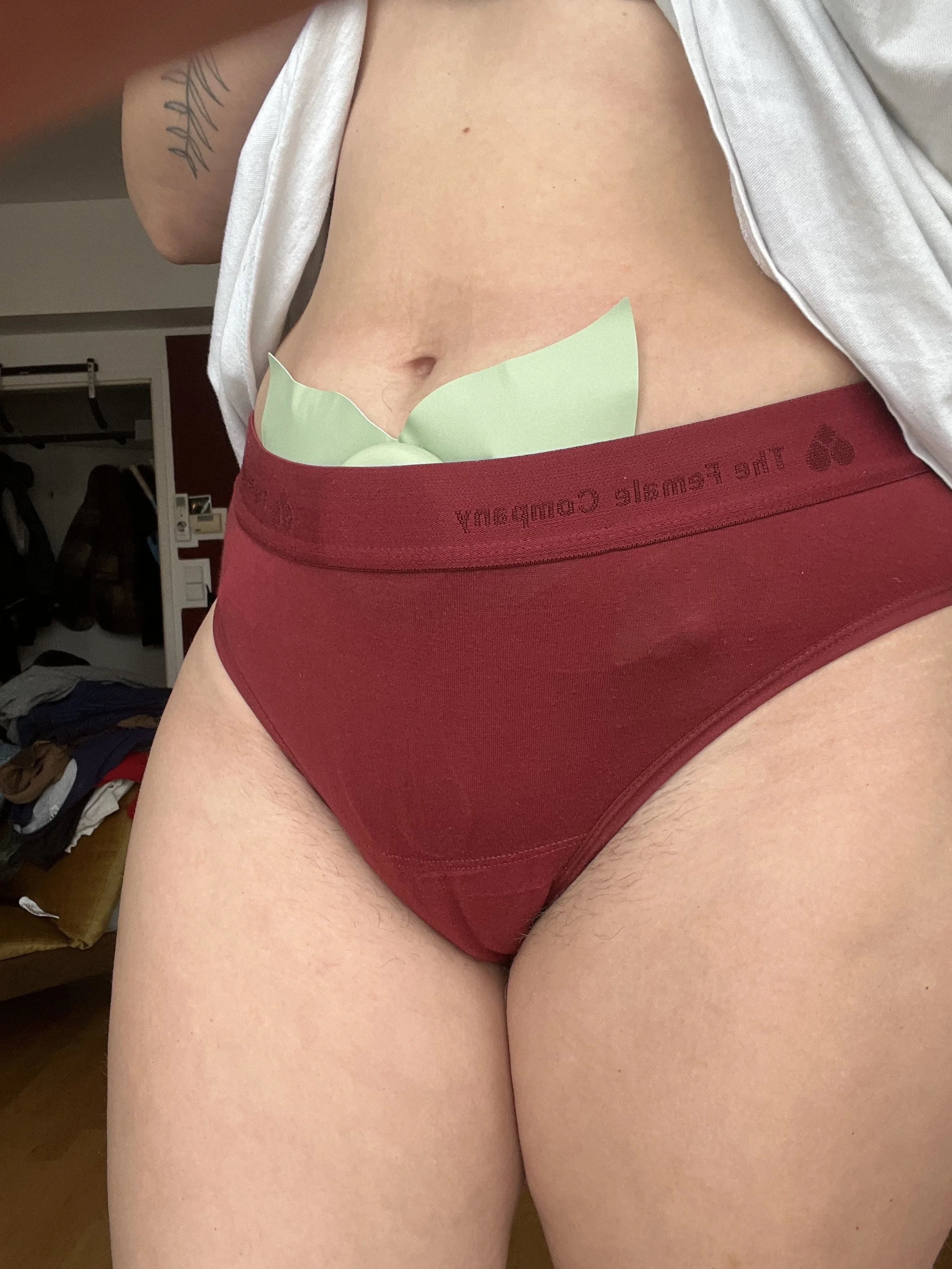 Close-up of a person wearing maroon underwear and a white top, with a torn green paper and a tattoo visible on the person's arm, in a room with a closet and clothes in the background.