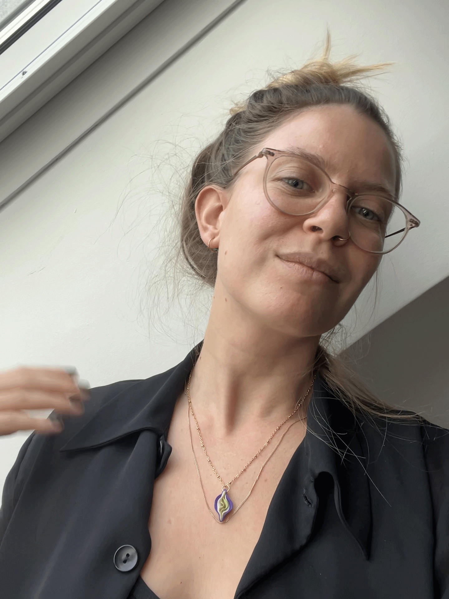 Eine junge Frau mit braunen Haaren, Brille und Halsketten, trägt eine schwarze Jacke und posiert vor einer weißen Wand.