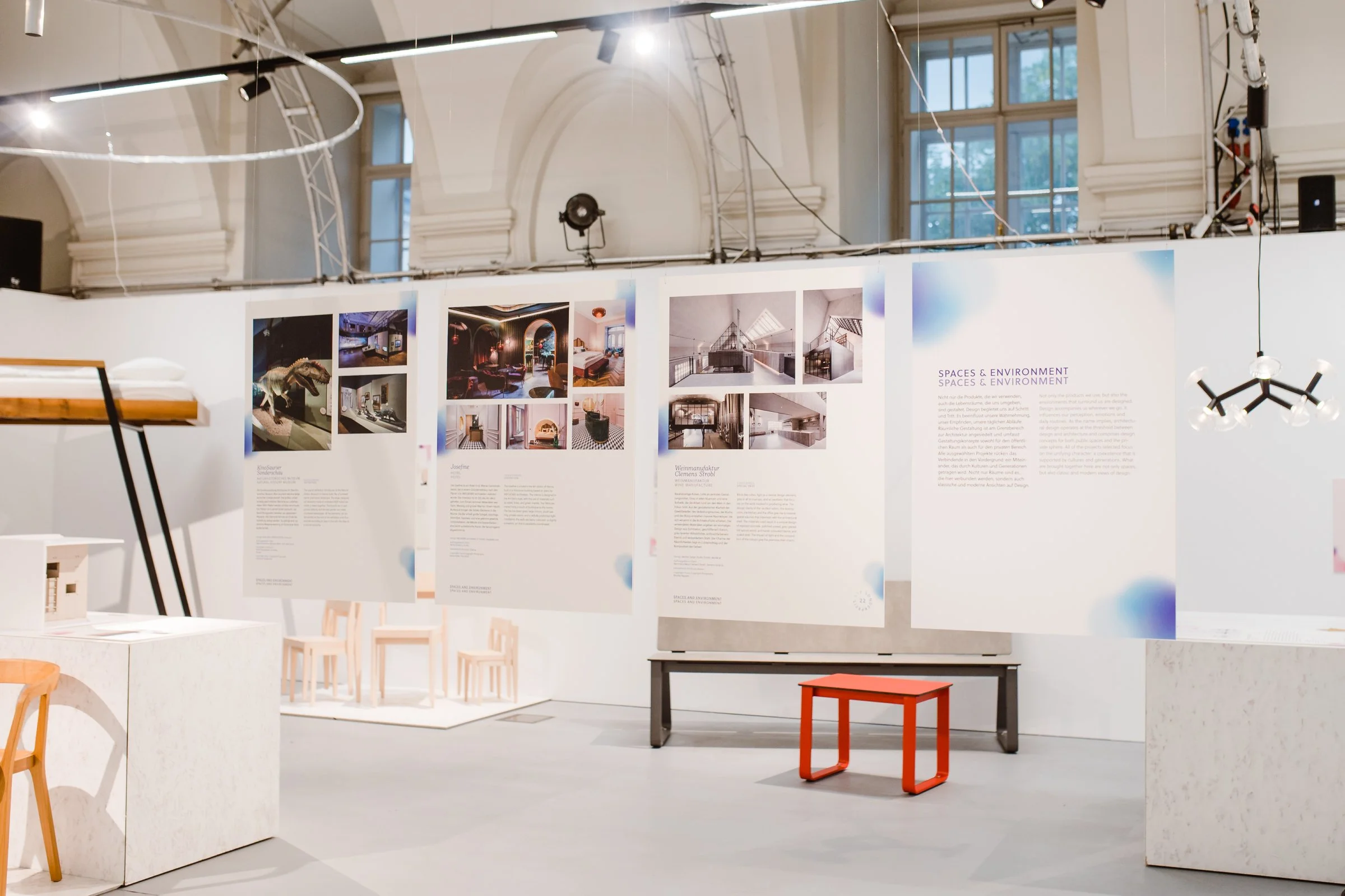 Ausstellung im Raum mit weißen Wänden, auf denen Fotos und Texttafeln zum Thema "Spaces & Environment" zu sehen sind. Im Hintergrund große Fenster, moderne Leuchten und technische Ausrüstung an der Decke.