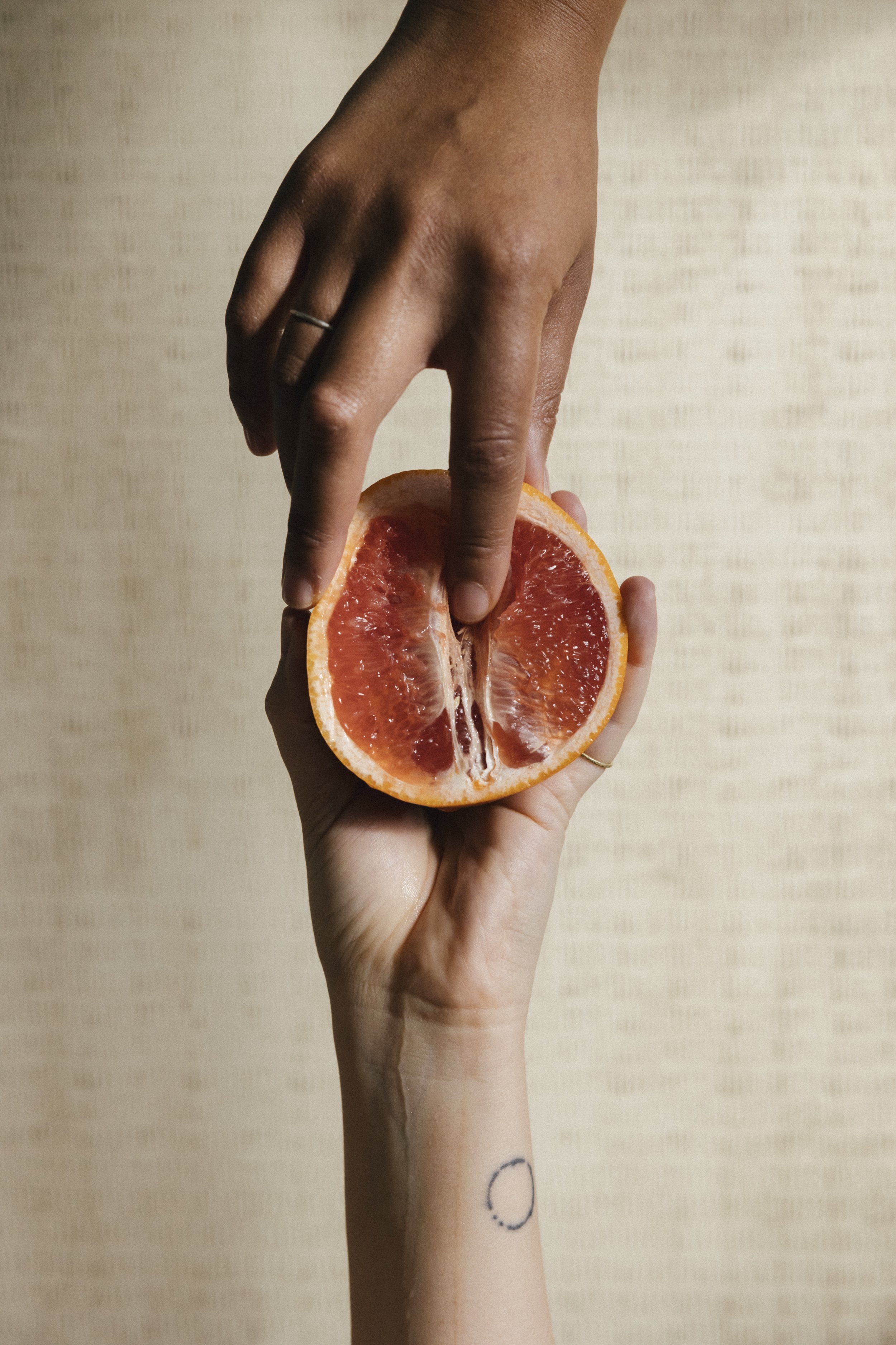 Zwei Hände, eine hält eine halbierte Grapefruit, die andere berührt die Frucht. Eine Hand hat einen Ring. Das Handgelenk einer Person hat eine kleine Tattoo mit einem Halbmond.