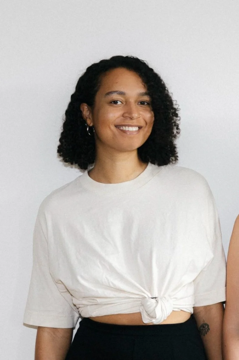 Eine lächelnde Frau mit schwarzen, lockigen Haaren trägt ein weißes T-Shirt mit Knoten vor dem Bauch, vor einem weißen Hintergrund.