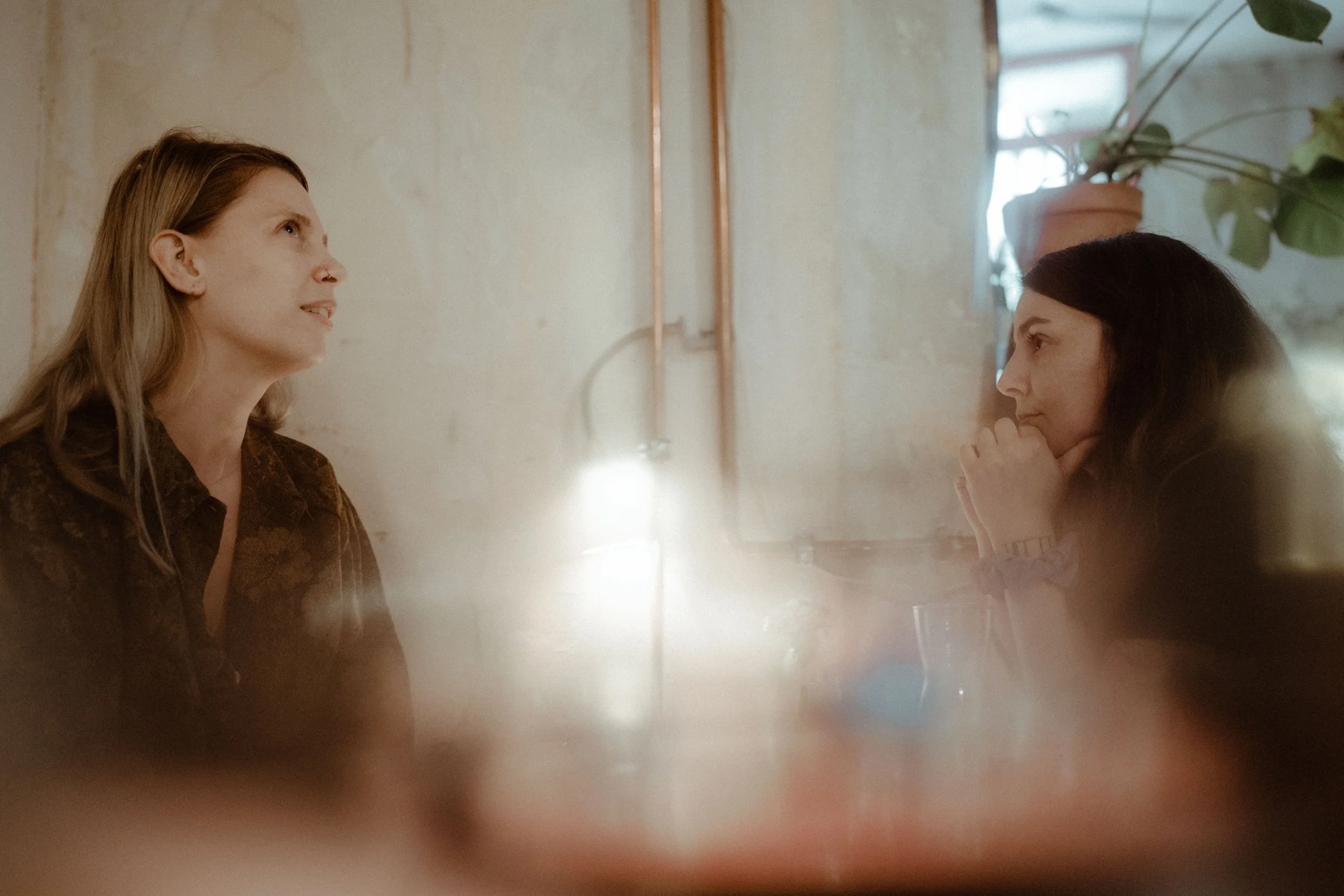 Two women sit across from each other at a table in a cozy, softly lit room with a rustic wall and potted plant in the background. The woman on the left has long light brown hair and is wearing a brown top. The woman on the right has long dark hair and rests her chin on her hand, appearing to listen attentively.