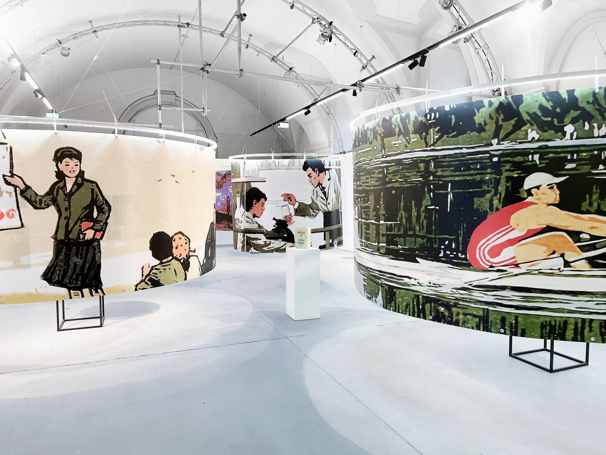 Ausstellungsraum mit großen runden Bannern, die comicartige Szenen mit Menschen zeigen, unter weißen Bögen und Beleuchtung.
