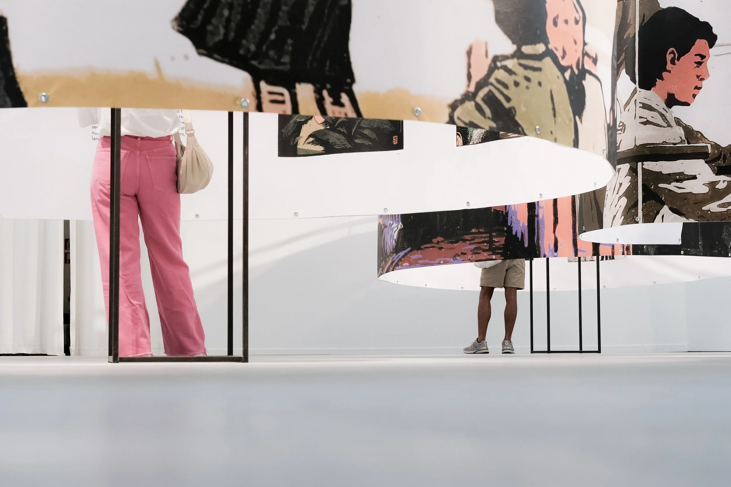 Menschen in einem Kunstmuseum betrachten große, bewegliche Wandkunstwerke mit abstrakten und porträtähnlichen Darstellungen, während die Szene im Ausstellungsraum minimalistisch gestaltet ist.