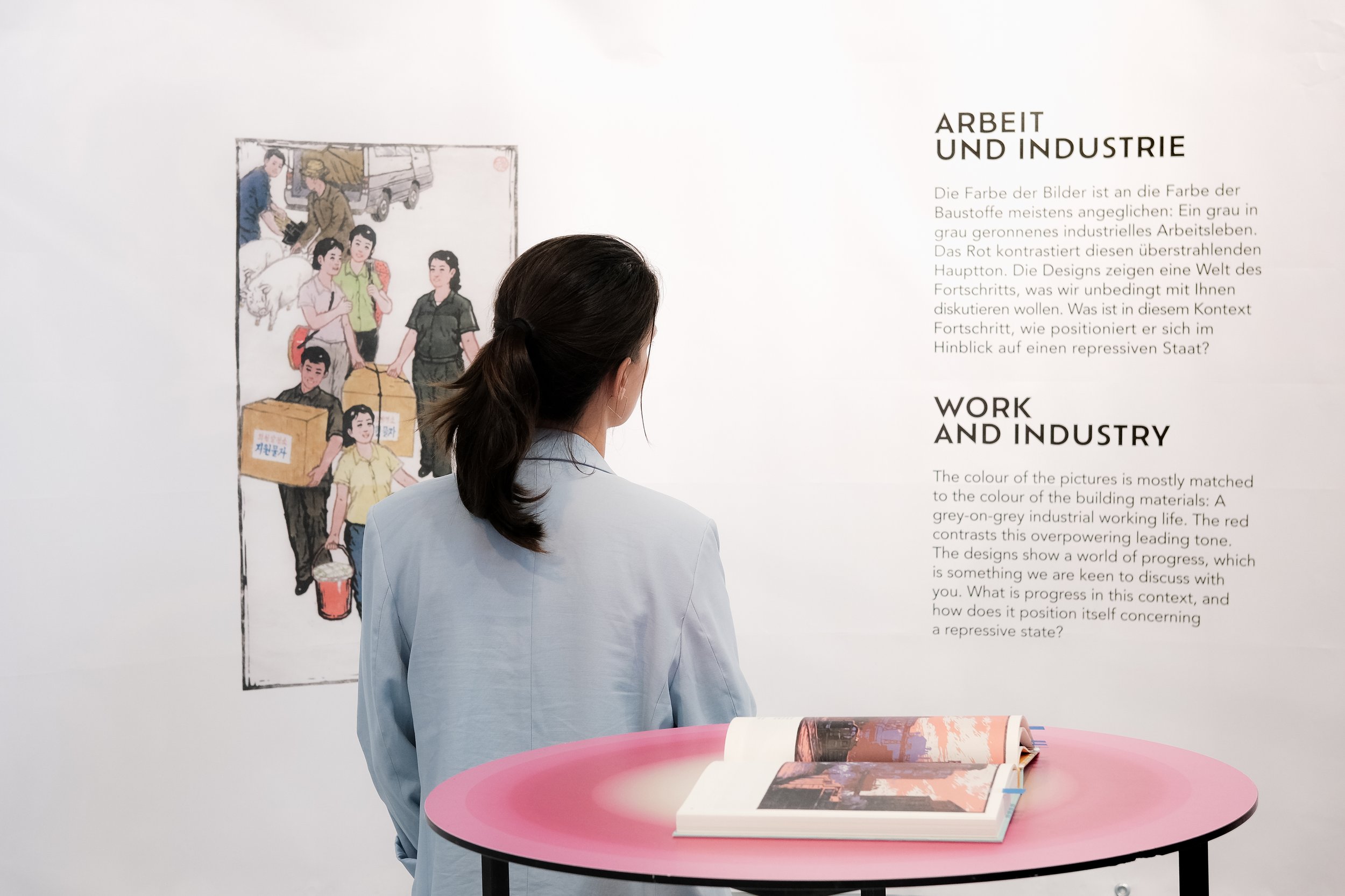 Eine Frau mit dunklen Haaren in einem hellblauen Blazer betrachtet ein Plakat an einer weißen Wand, das über Arbeit und Industrie spricht.