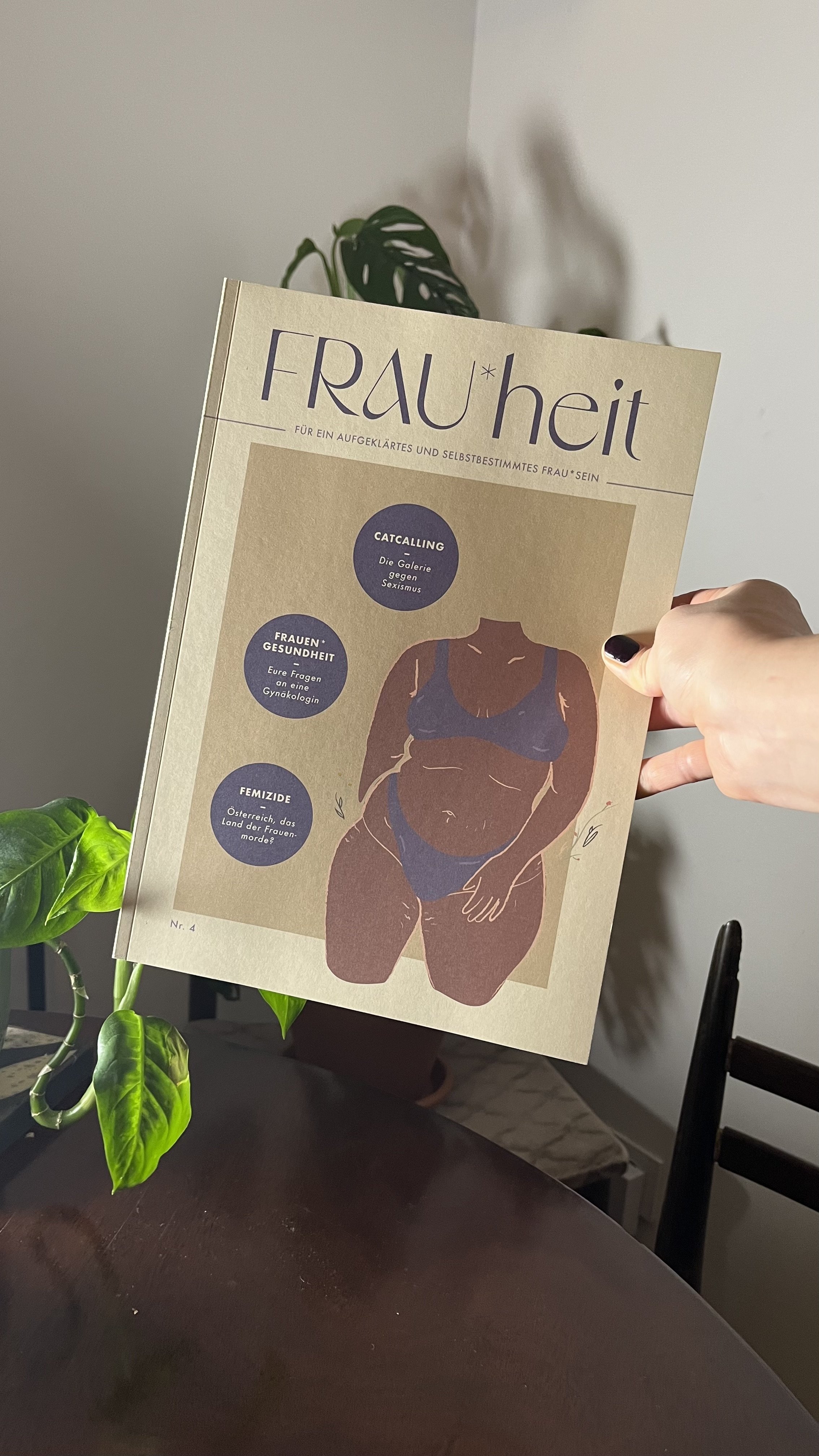 A person holding a magazine titled 'FRAU*heit' with an illustration of a woman in underwear on the cover, placed on a table near a green plant.