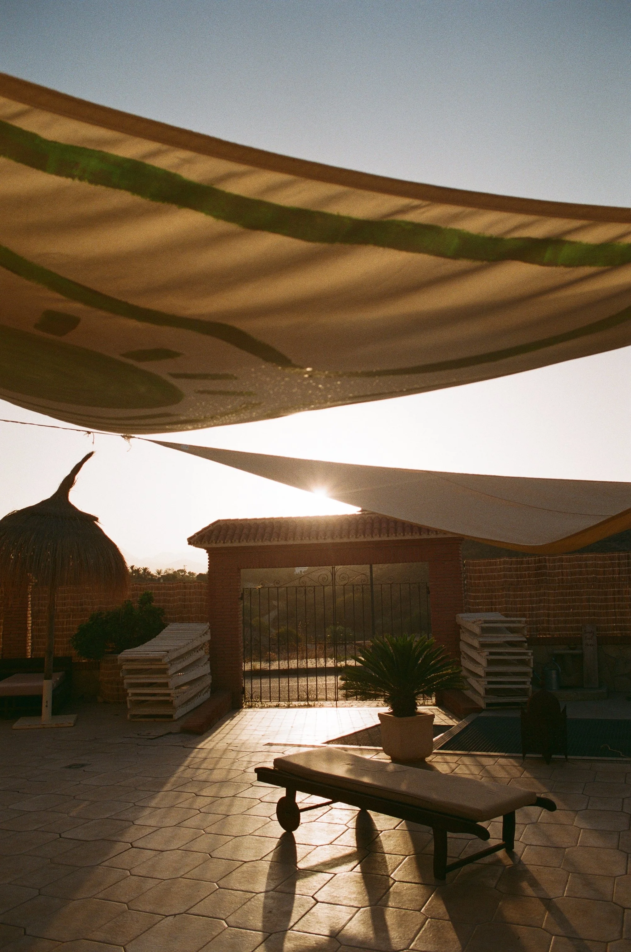 Ein Terrassenbereich mit gepflastertem Boden, einem Sonnenstuhl, einem Sonnenschirm und Töpfen mit Pflanzen, im Hintergrund eine Gartenmauer und das offene Tor, unter einem Sonnenschutzdach, bei Sonnenuntergang.