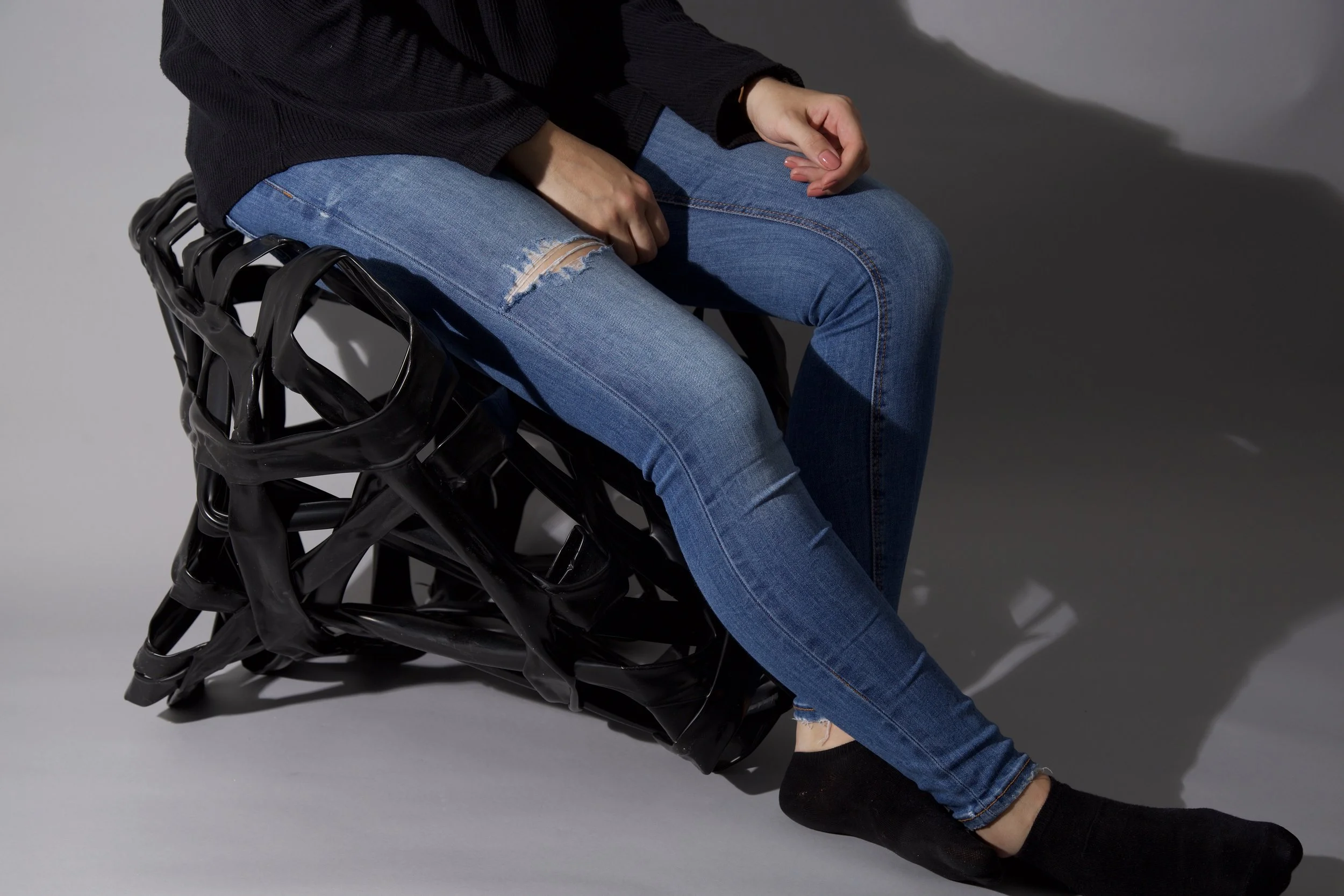 Eine Person sitzt auf einem schwarzen Stuhl aus Kunststoff, trägt eine schwarze Jacke, blaue Jeans mit Riss im Knie, schwarze Socken und Schwarzschuhe. Der Hintergrund ist grau, und das Foto ist seitlich aufgenommen.