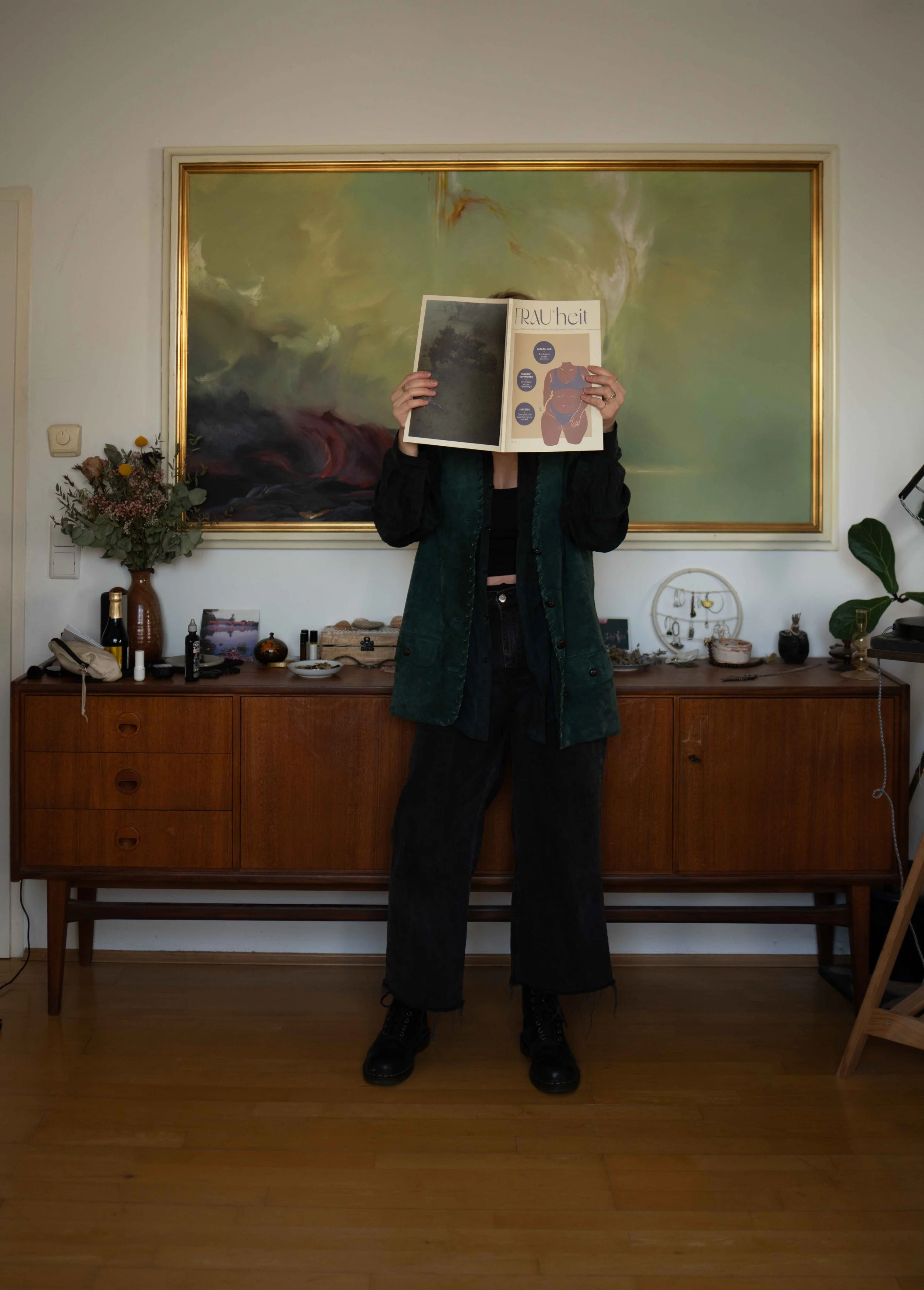 Person steht vor einem Holzmöbel, Foto wird durch aufgeklautes Magazin verdeckt, im Hintergrund ein großes abstraktes Gemälde an der Wand.
