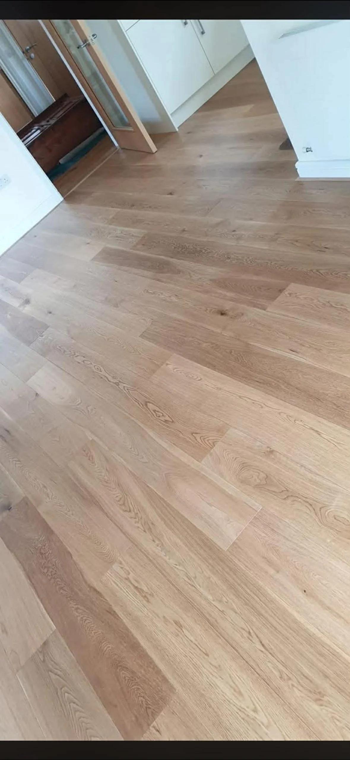 Photo of a hardwood floor in a room with white walls. Visible door frames and a partially open wooden door are in the background.
