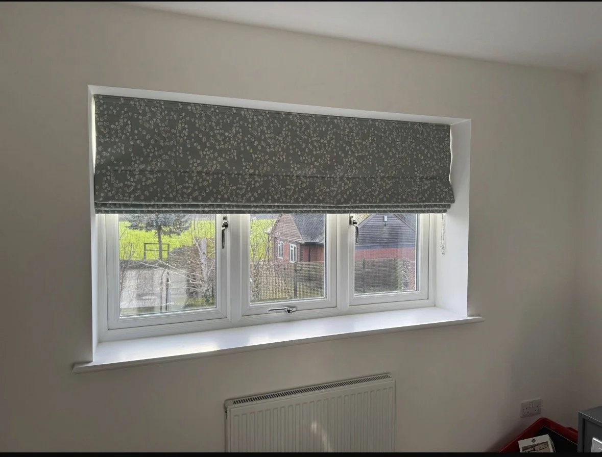 A white-framed window with a floral-patterned gray Roman blind partially covering it, set in a white wall with a radiator beneath. Outside the window, trees, houses, and a fence are visible.