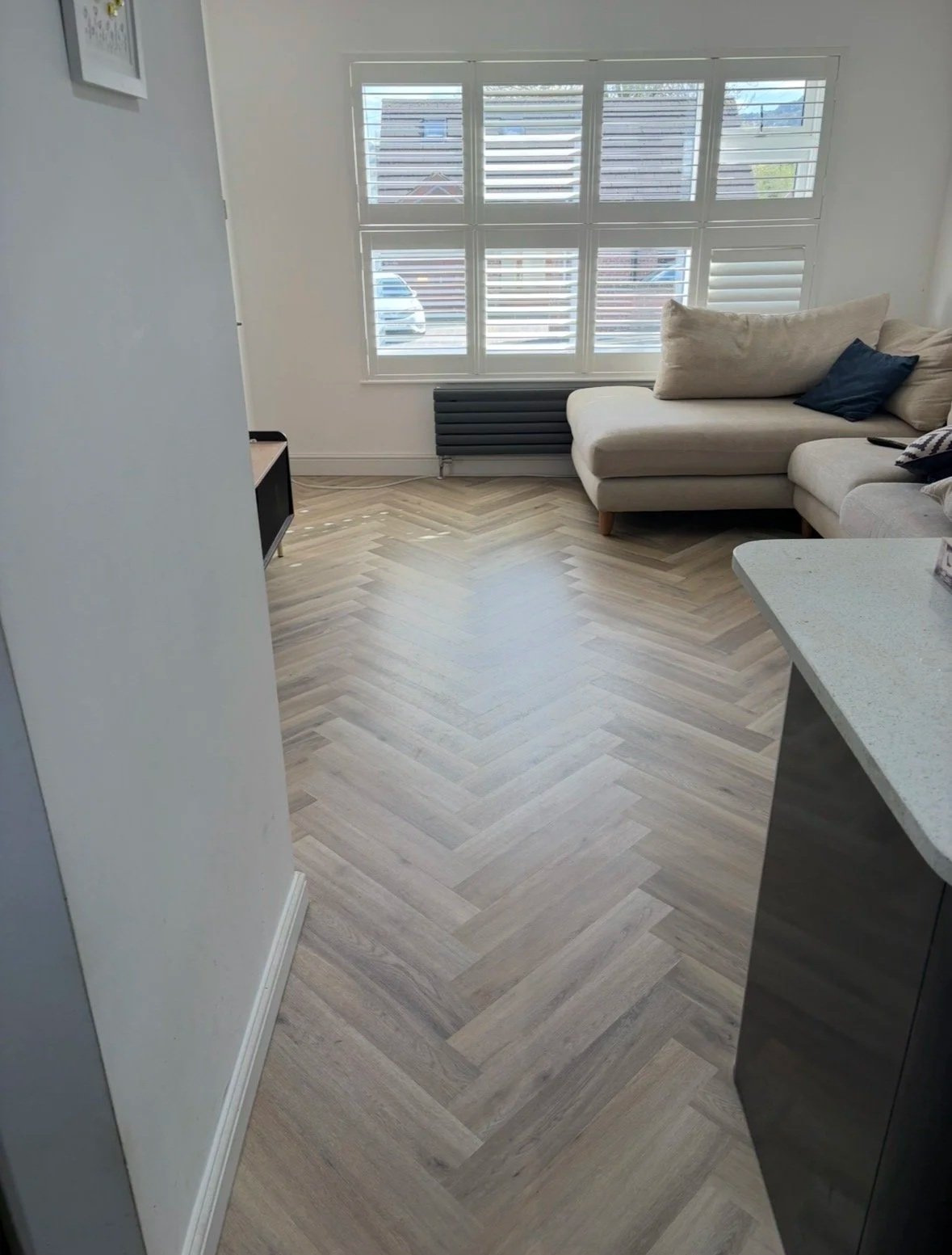 Living room with a large window, beige L-shaped sofa with pillows, small black side table, radiator, and light-colored herringbone wood floor.