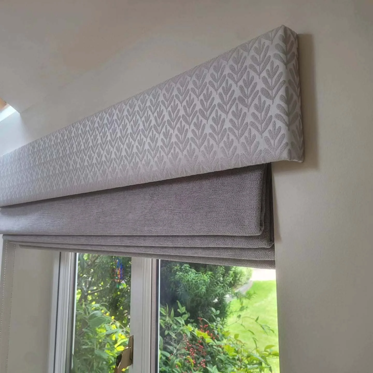 A window with a partially drawn beige curtain and a landscape view of green trees and grass outside.