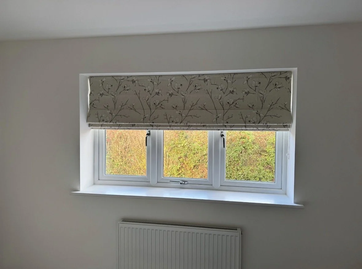 A window with three panes and a floral-patterned roller blind in a room with white walls and a radiator below the window.