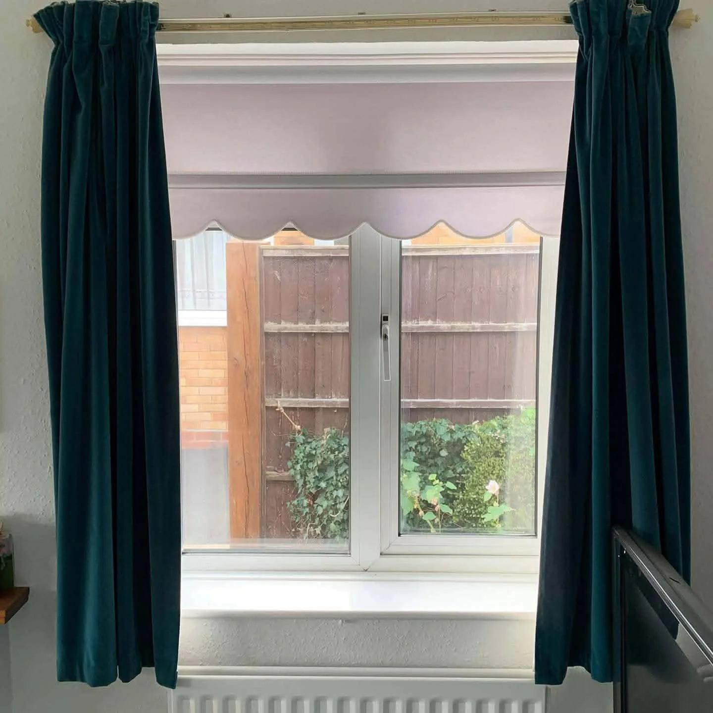View through a window with blue curtains showing a wooden fence, greenery, and part of a brick wall outside.