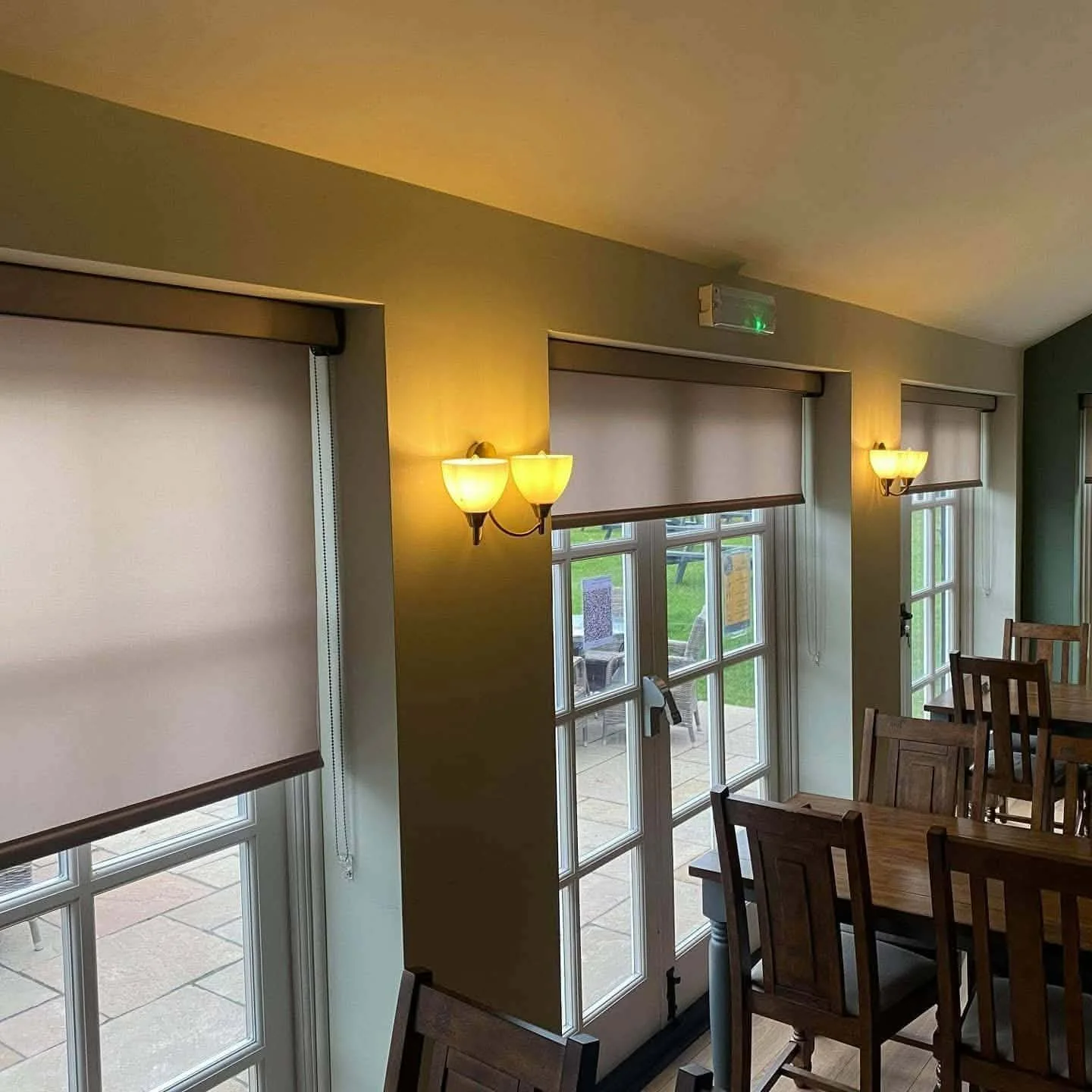 Interior of a dining area with wooden chairs and tables, several large windows with blinds, and wall-mounted light fixtures.