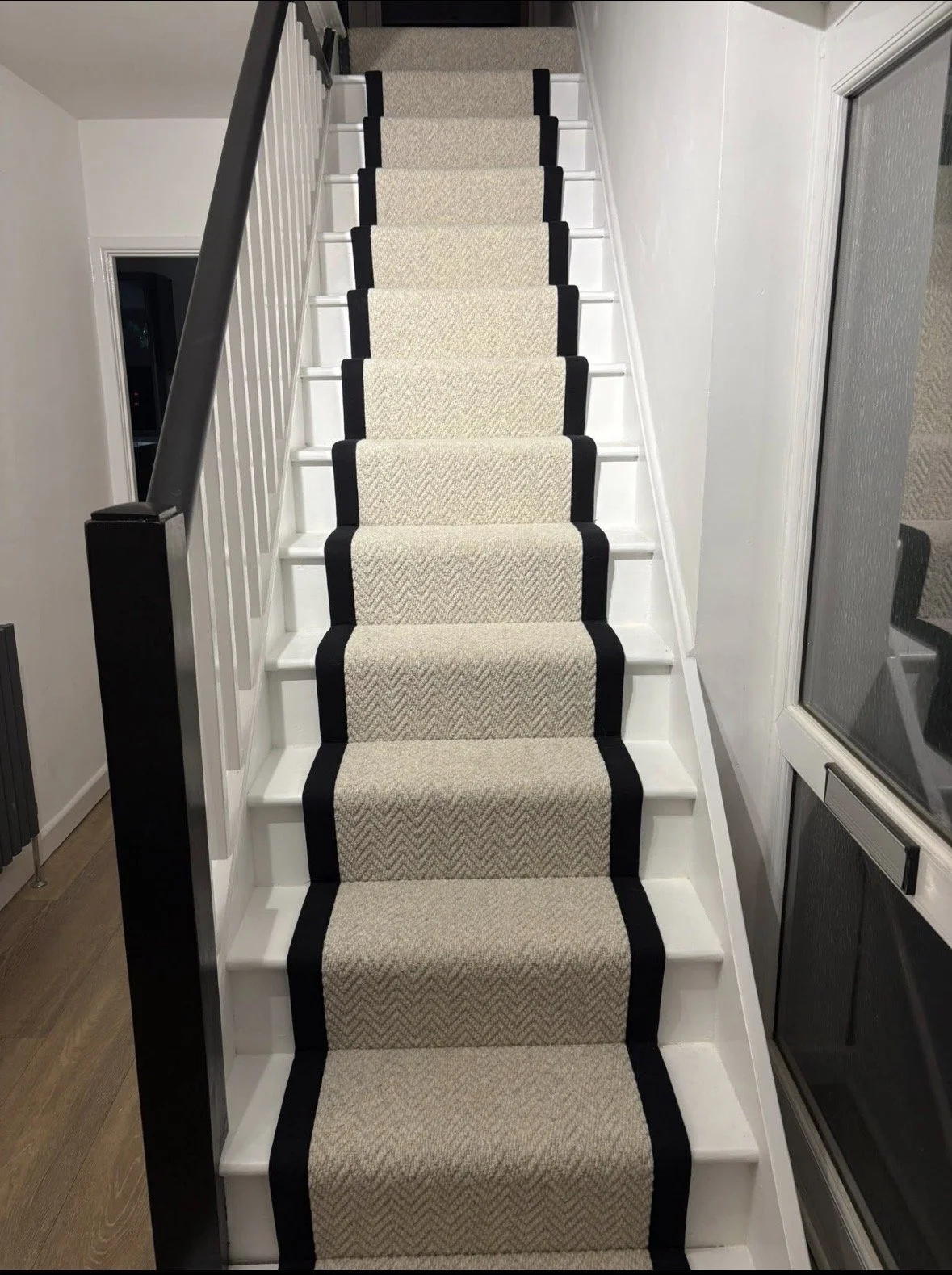 White staircase with beige runner carpet bordered in black, leading upstairs, with a black handrail on the left and a glass door on the right.
