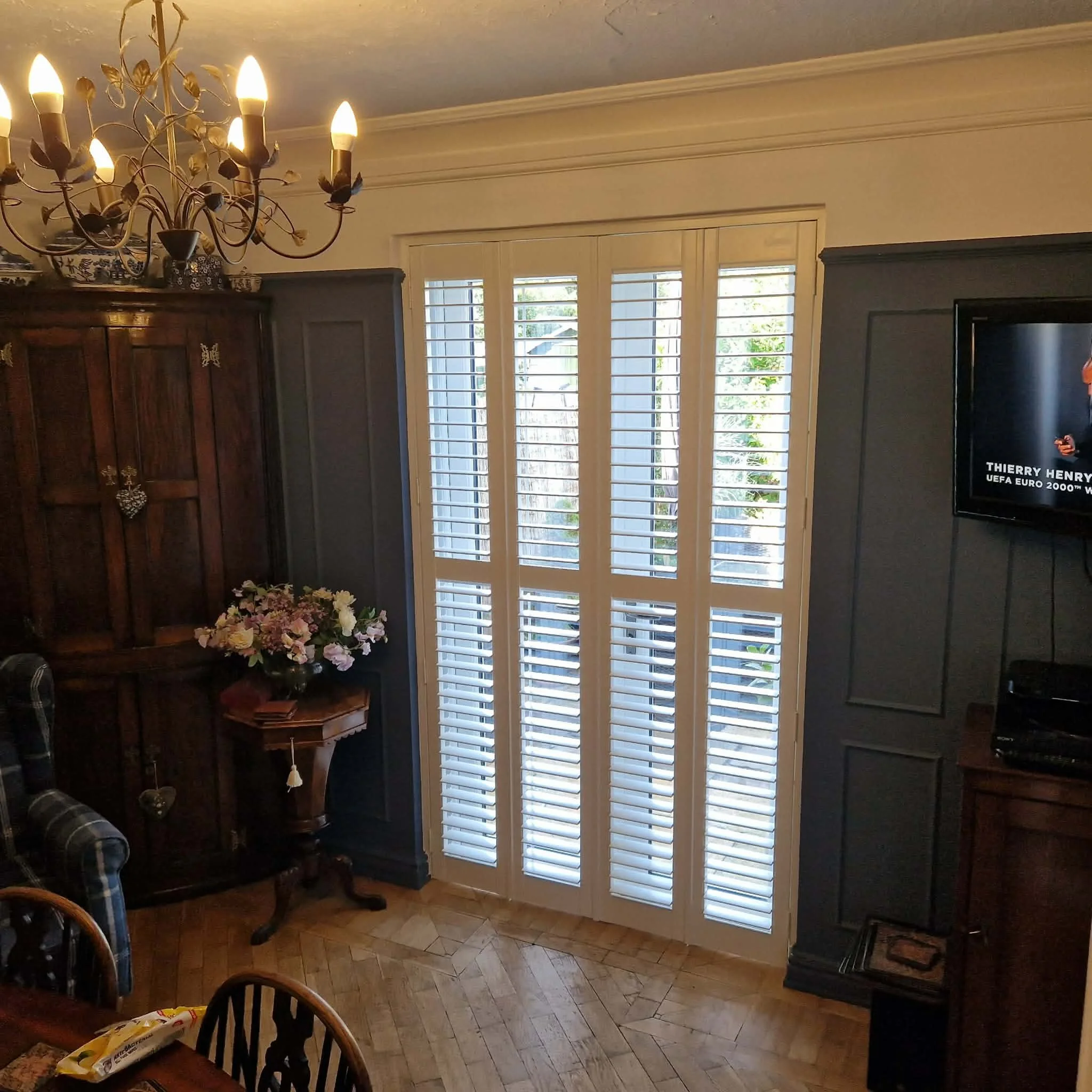 Living room with a chandelier, large window with plantation shutters, dark wood cabinet, floral bouquet on a small table, and a television with a football match playing.
