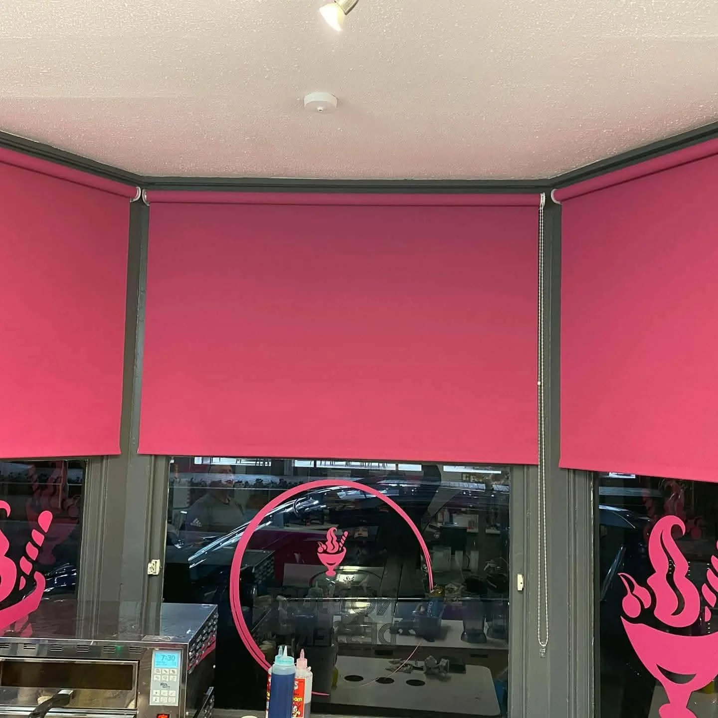 Pink window shades seen in a storefront, with a glass door and window below. The window features a pink flamingo signage. Inside are some condiment bottles and a microwave oven, with cars visible outside through the glass.