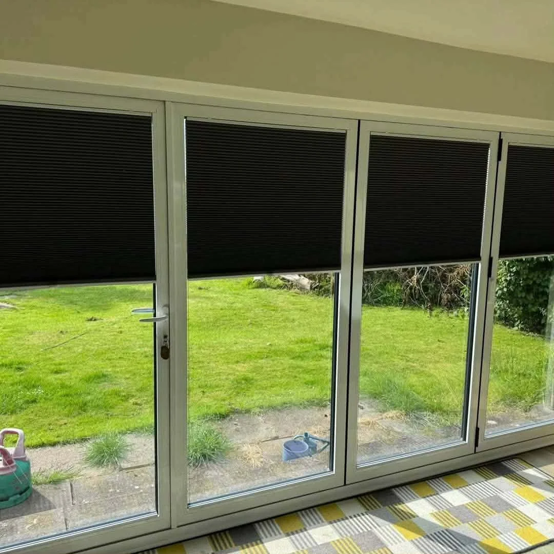 View of a sliding glass door with partially closed black blinds, opening to a green backyard with grass, plants, and garden tools outside.