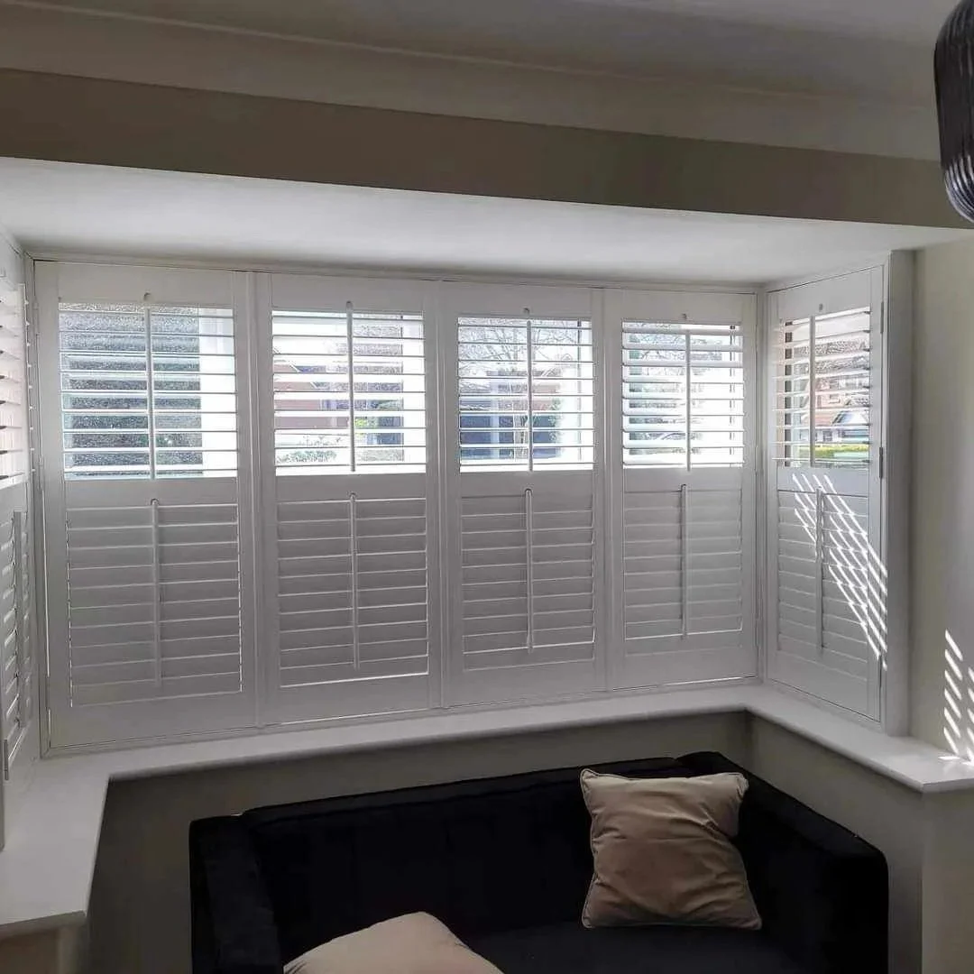 Bright bay window with white plantation shutters, black couch with beige pillows, and sunlight casting shadows inside a living room.