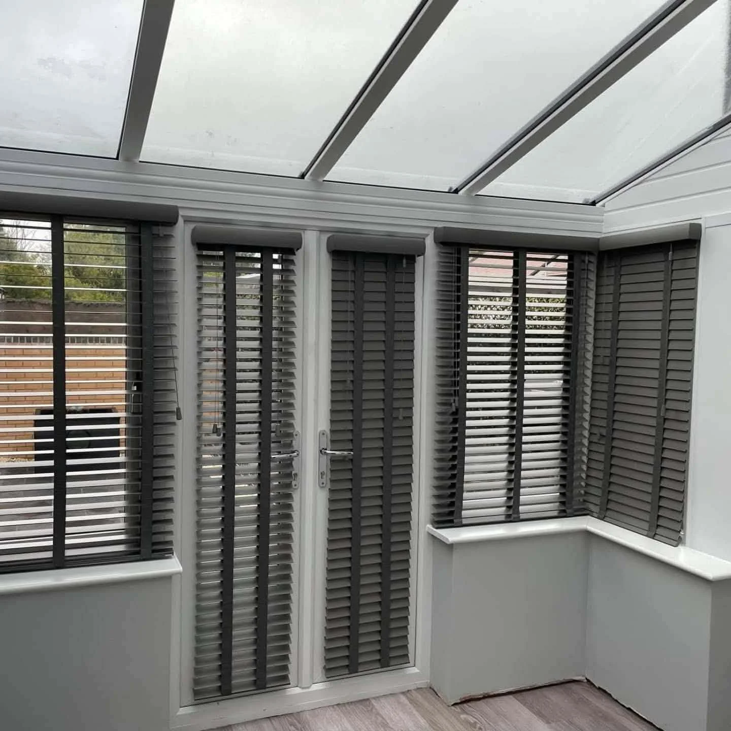 Indoor enclosed space with white framing, grey plantation shutters on windows, glass roof, and light-colored wooden floor.