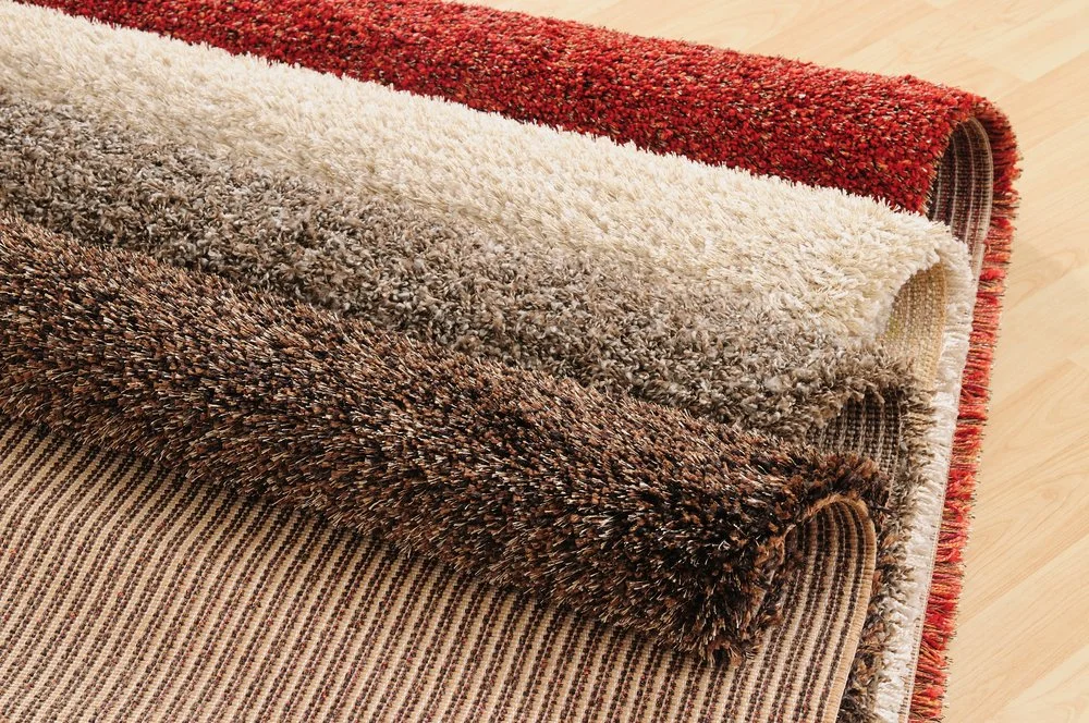 Close-up of three rolled and layered area rugs with textured patterns in shades of brown, beige, cream, and red, on a light wooden floor.