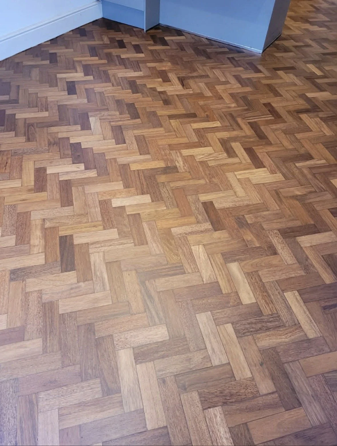 Close-up of a hardwood parquet floor with a herringbone pattern, wood planks in varying shades of brown, underneath a corner of light blue wall and white baseboard.
