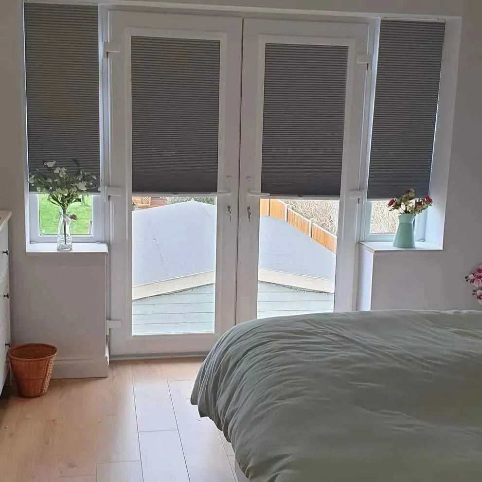 A bedroom with a large window and a glass door, covered by dark gray blinds. Two small windows on either side hold flower vases, one with white flowers and another with red and white flowers. Part of a bed with a light-colored comforter is visible in the foreground.