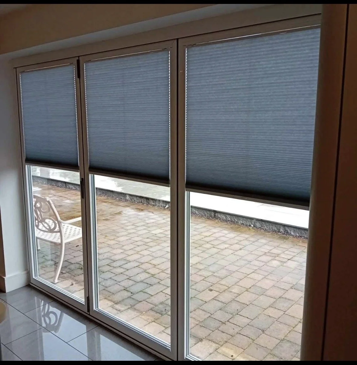 Sliding glass doors with closed blue window blinds, looking out onto a brick patio with a white metal outdoor bench.
