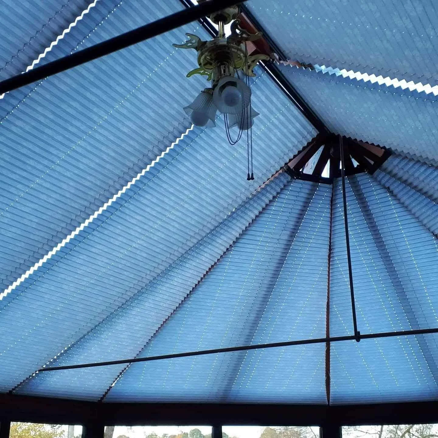 Interior view of a building ceiling with an ornate chandelier hanging from the center, featuring multiple glass lamps and decorative metalwork, surrounded by a blue corrugated metal roof with neon strip lighting.