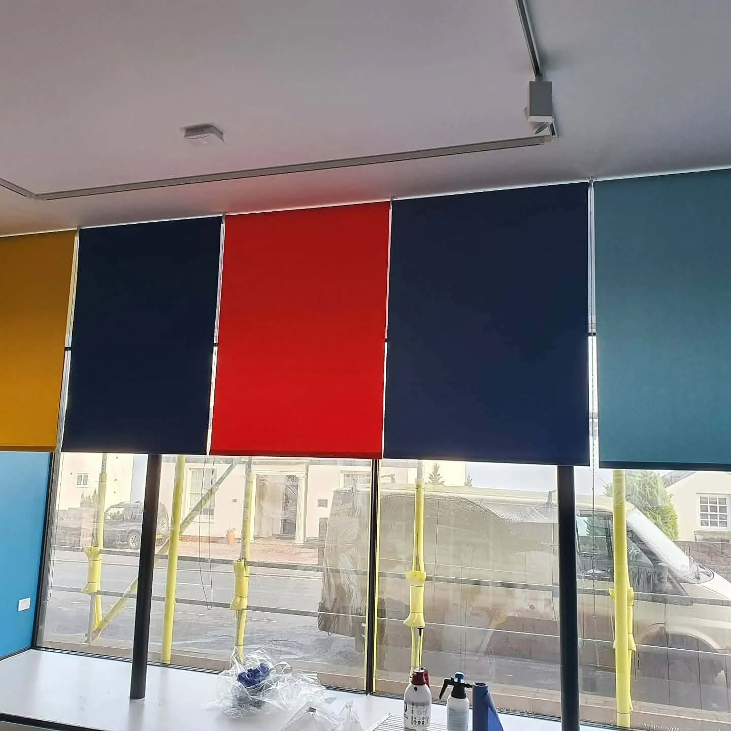 Indoor space with large colored fabric panels hanging from the ceiling in yellow, navy blue, red, navy blue, and teal. The panels are in front of large windows showing a parking lot and buildings outside. There are tools and cleaning supplies on the white counter below the panels.