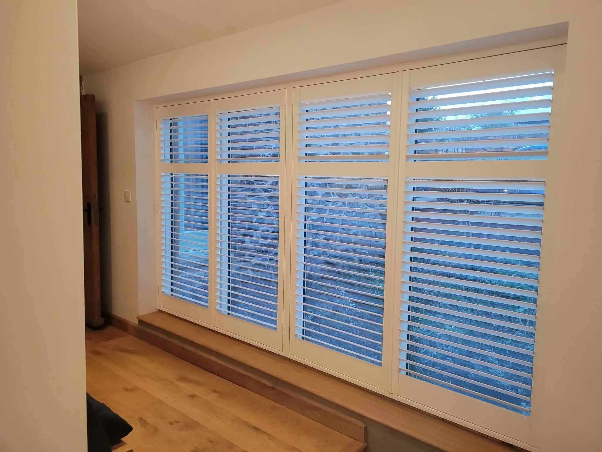 Interior of a room with large white-framed windows and wooden shutters, showing a view of trees outside at dusk.