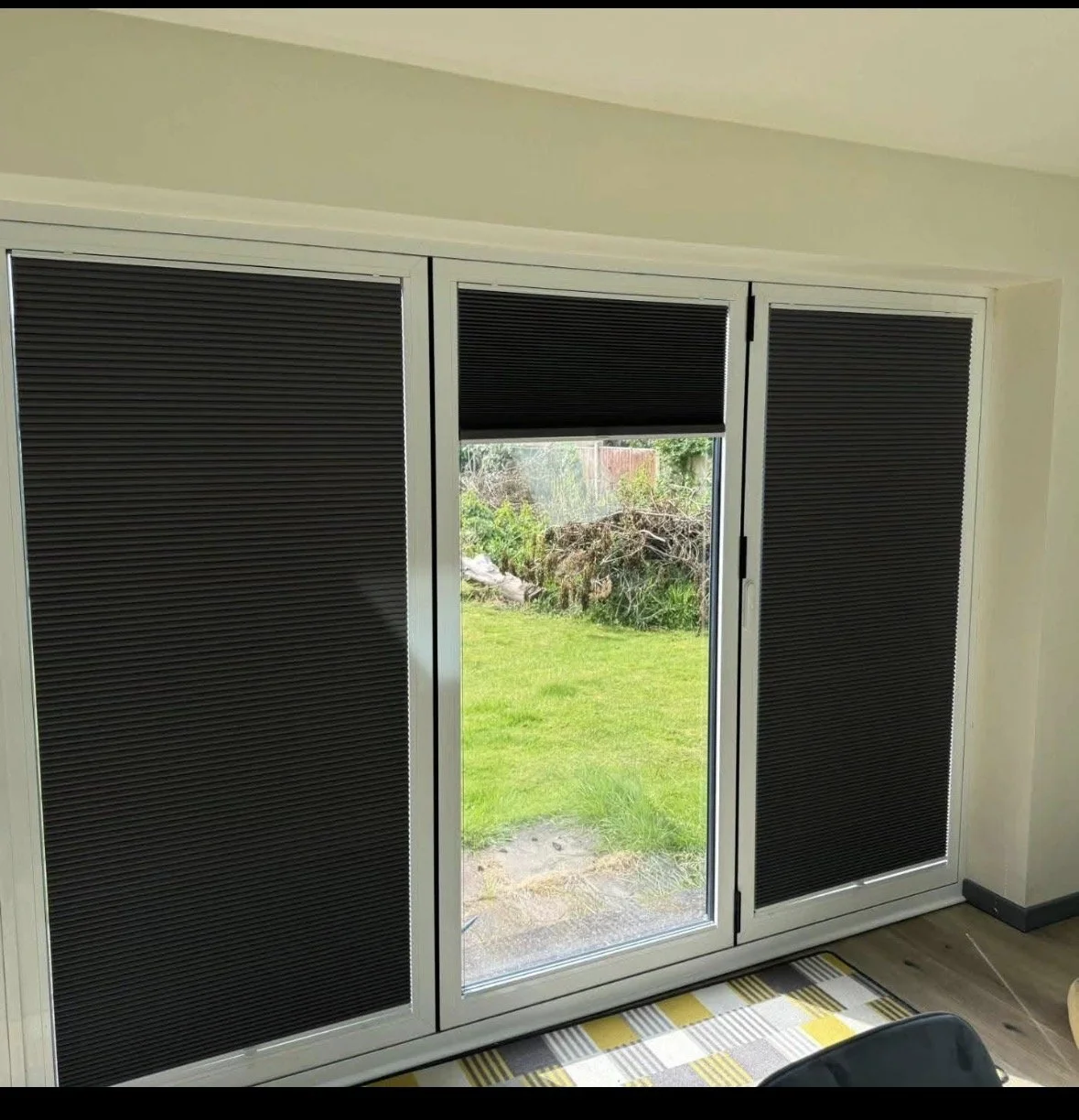 Sliding glass door with black blinds partially covering the door, leading out to a grassy backyard with bushes and trees.