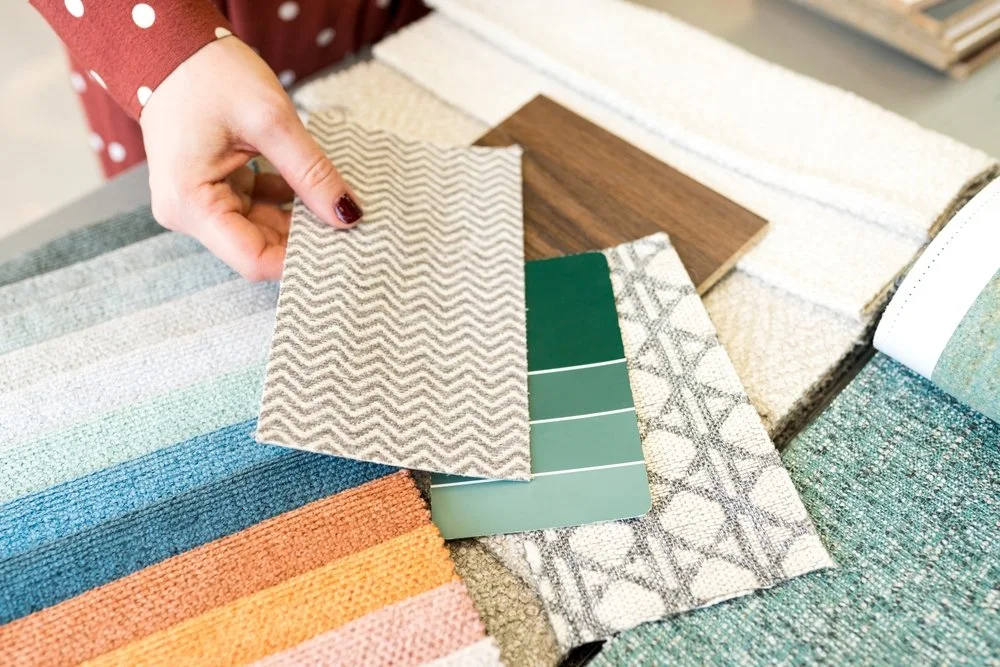 A person with red nail polish holding a small fabric sample with a zigzag pattern over an assortment of fabric and material swatches, including wood and textured fabrics.