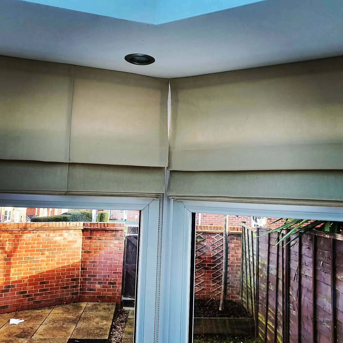 View of an outdoor patio area with a brick wall, concrete floor, and a part of a small garden with plants, seen through a glass door from inside a room with beige window blinds and a ceiling light.