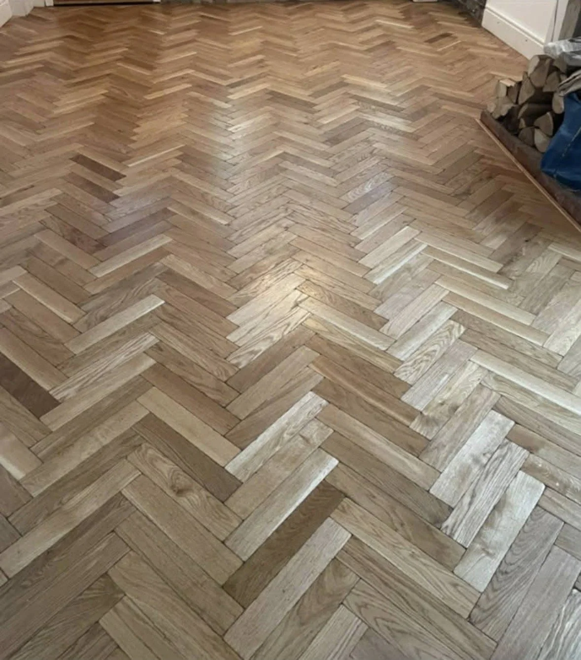 Wooden parquet flooring arranged in a herringbone pattern in a room with a corner of a wood stack visible.