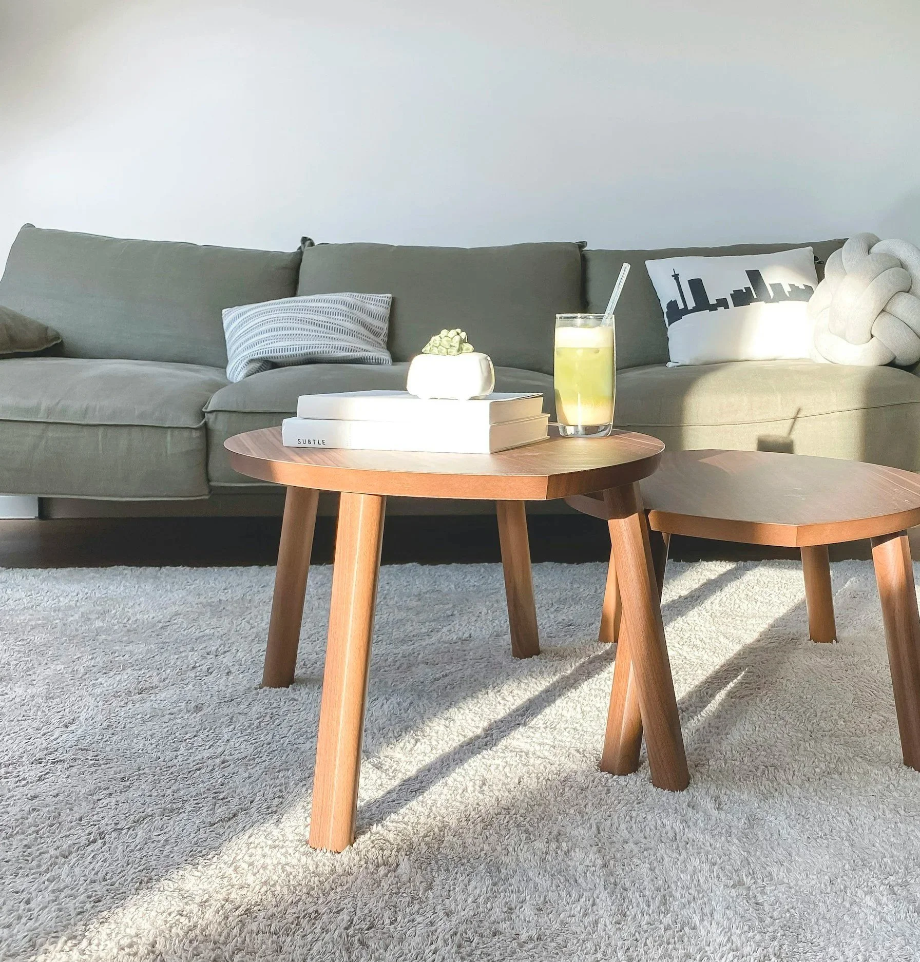 Living room with a beige sofa, decorative pillows, a round wooden coffee table, a white plush knot pillow, a glass of lemonade with a straw, a small white vase with green decorative objects, books, and a textured white rug.