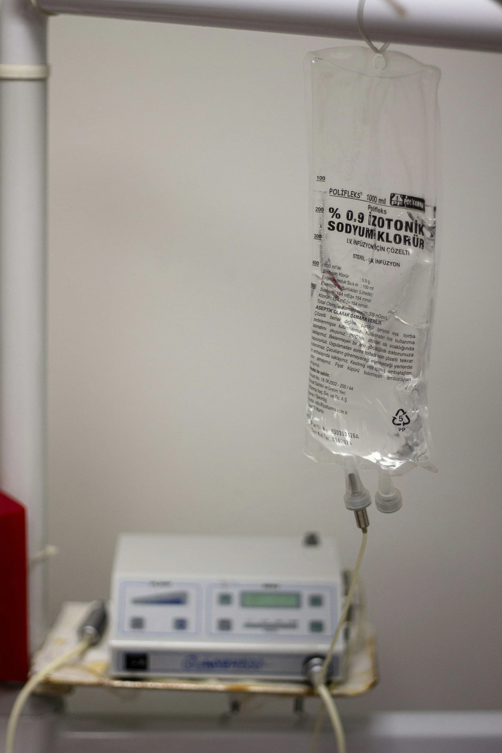 A IV infusion bag hanging next to a medical infusion pump on a hospital bed, with the bag containing a clear liquid and the pump connected via tubes.