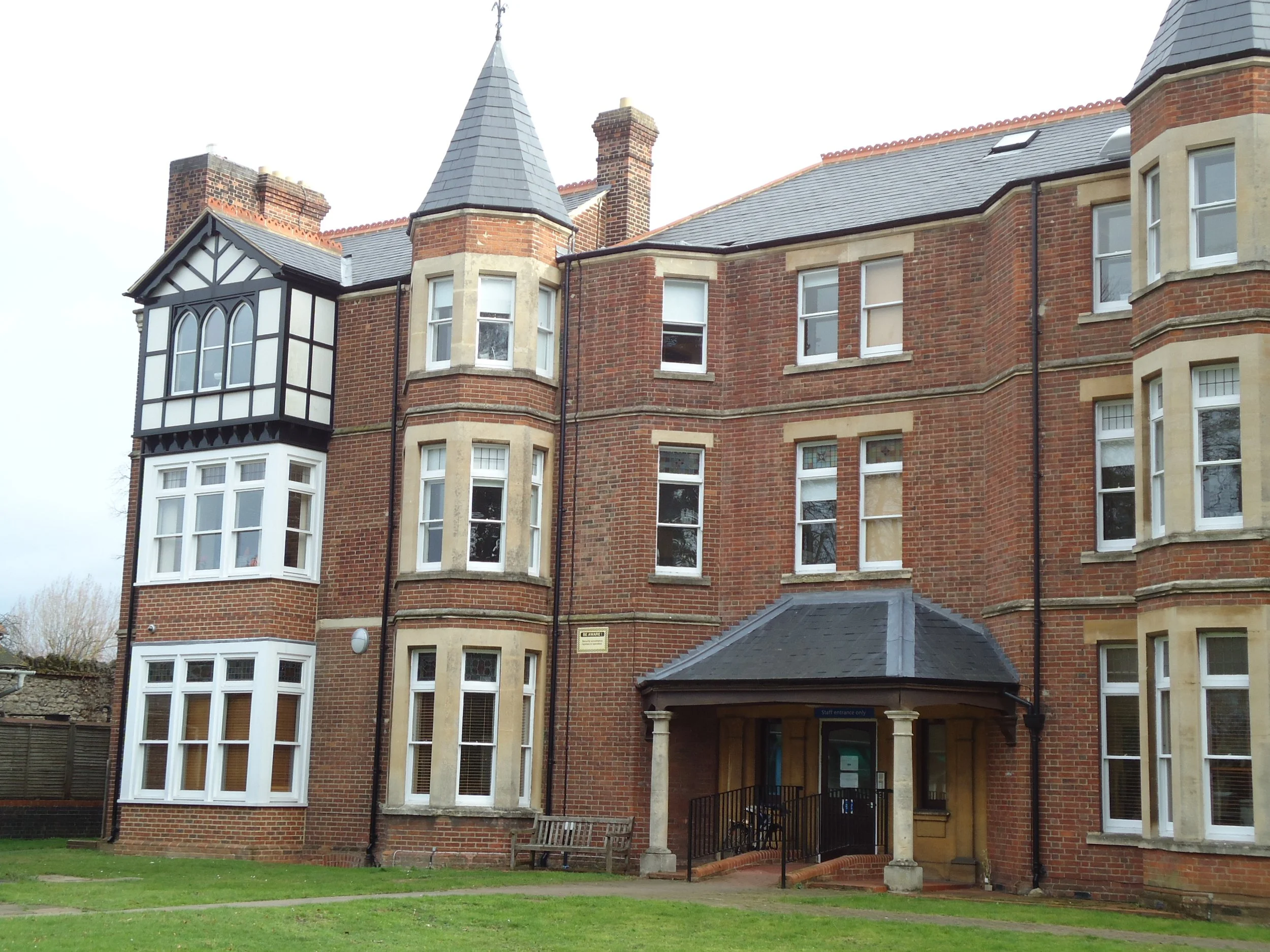 Red brick multi-story apartment building with bay windows, turret-like corner structures with pointed roofs, and a covered entrance with columns, surrounded by a grassy area.