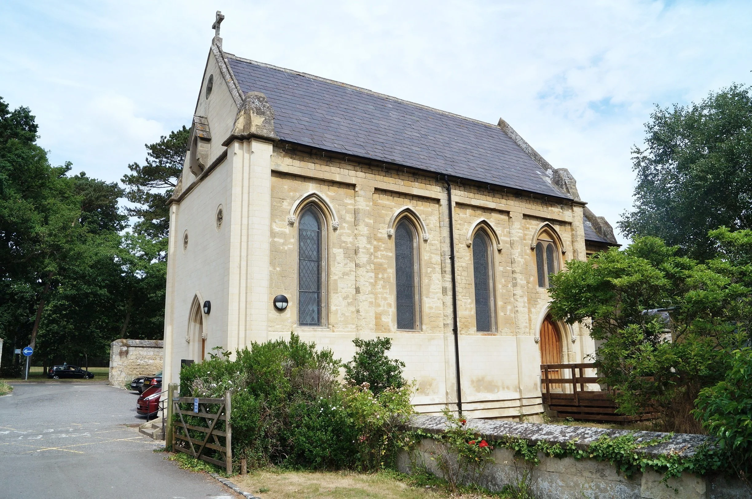A small historic church with Gothic windows, surrounded by greenery and parked cars, under a partly cloudy sky.