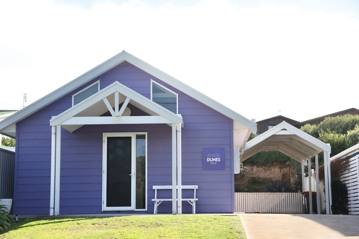 A small purple beach house with white trim and a white bench in front, featuring a sign that reads 'Dunes Property' on the side, and a carport to the right.