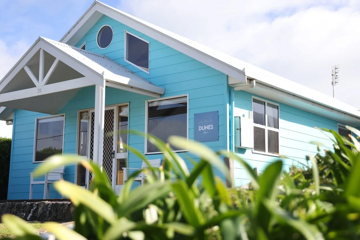 A bright blue house with white trim, a porch, and several windows, located in a coastal area called Dunes.