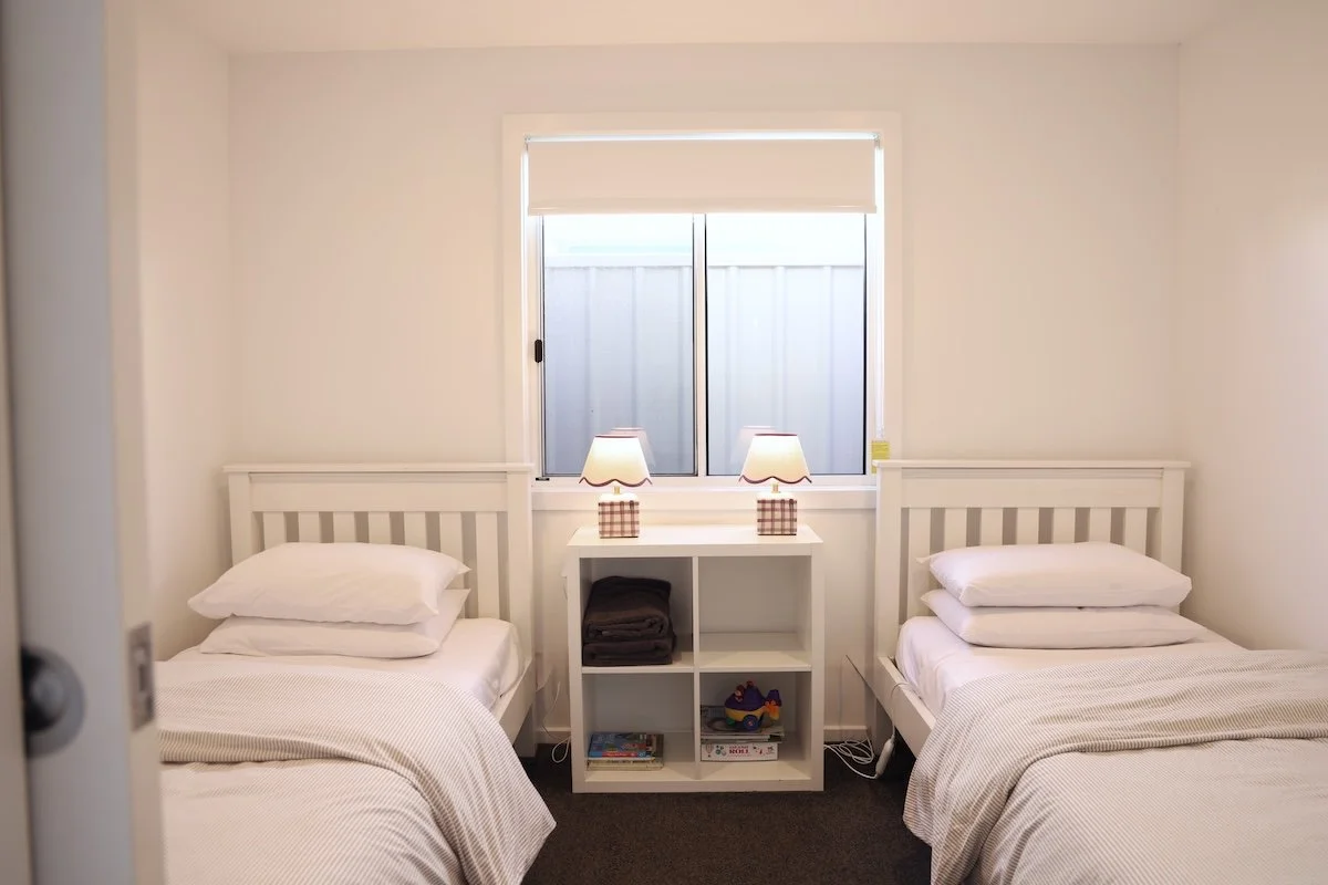 A bedroom with two single beds with white bedding and pillows, a white shelf in between with two lamps and books, against a wall with a window.