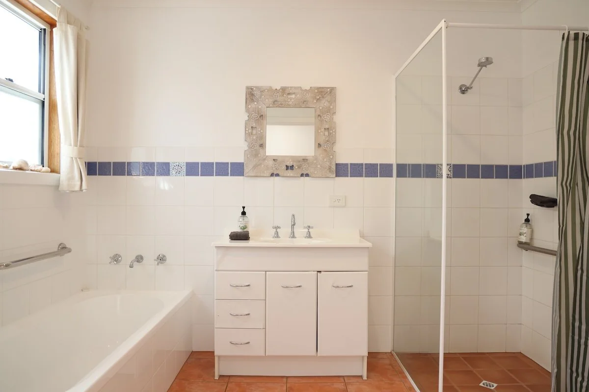 A bathroom featuring a bathtub on the left, a small white vanity with a mirror above it in the center, and a walk-in shower on the right. The bathroom has white tiled walls with a blue decorative border, a window with curtains, and brown tile floorin