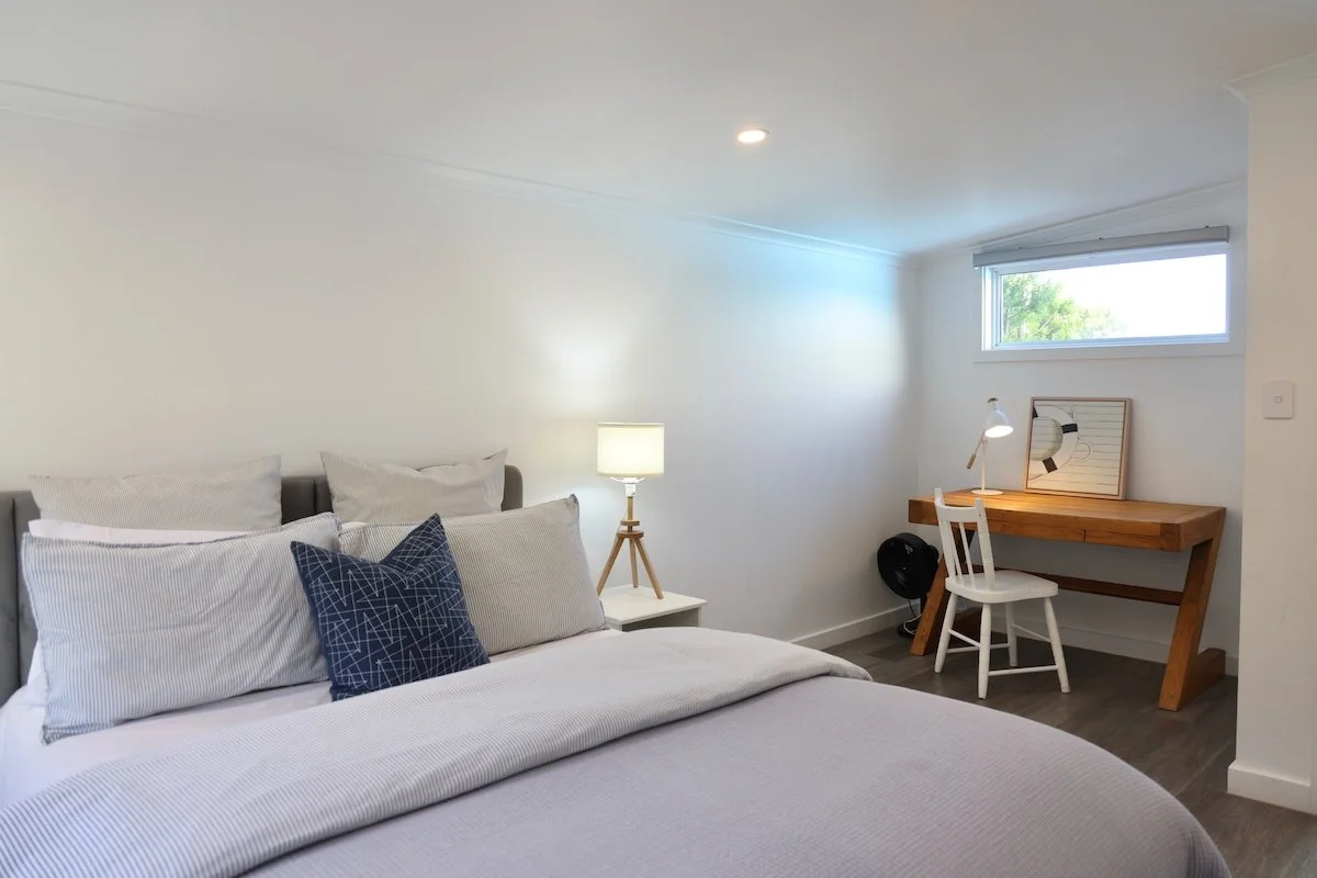 A bedroom with a large bed covered in white and gray pillows, a small white nightstand with a lamp, and a wooden desk with a white chair and a framed artwork.