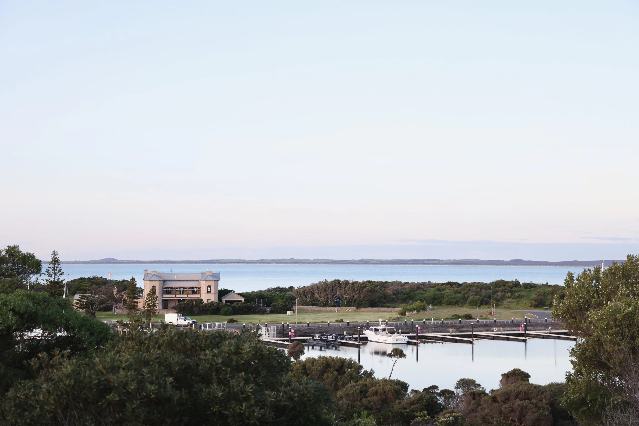 A scenic view of a lakeside area with a house, trees, and boats docked at a marina, with a body of water and distant land in the background under a clear sky.