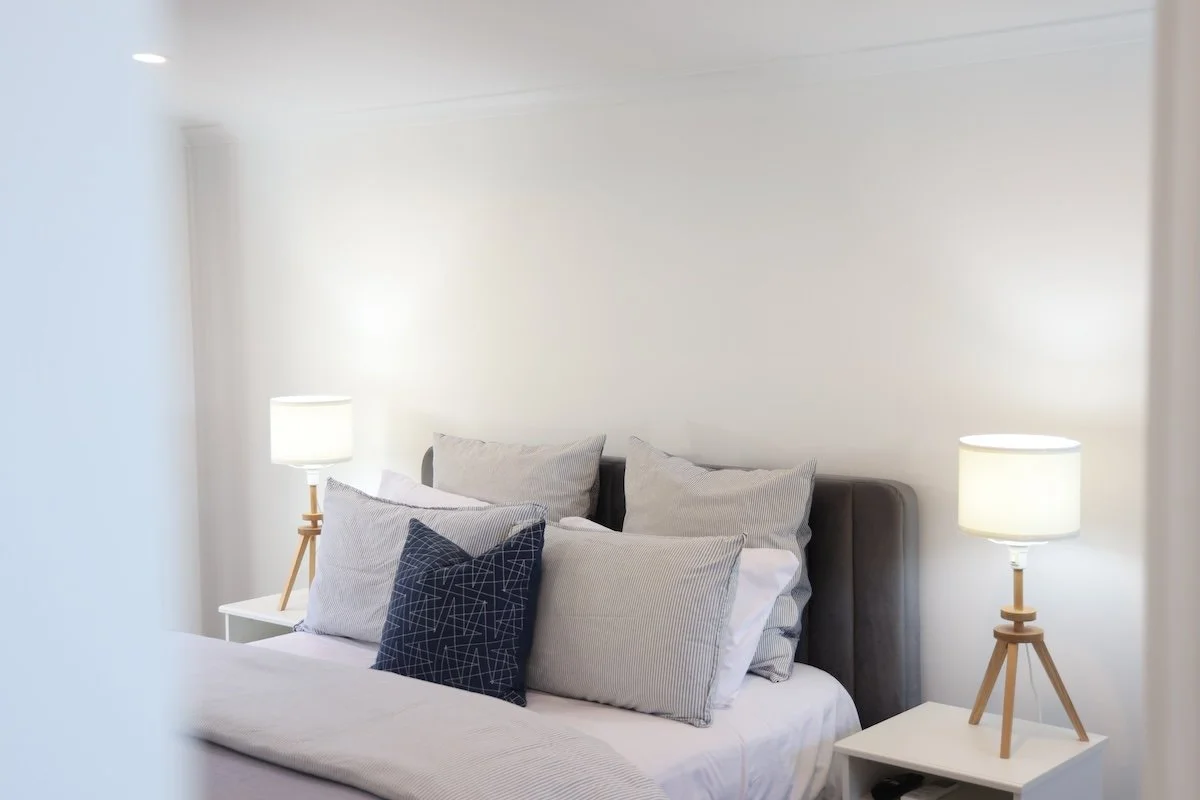Neatly made bed with gray and white pillows, flanked by white nightstands with modern bamboo tripod lamps in a minimalist bedroom.