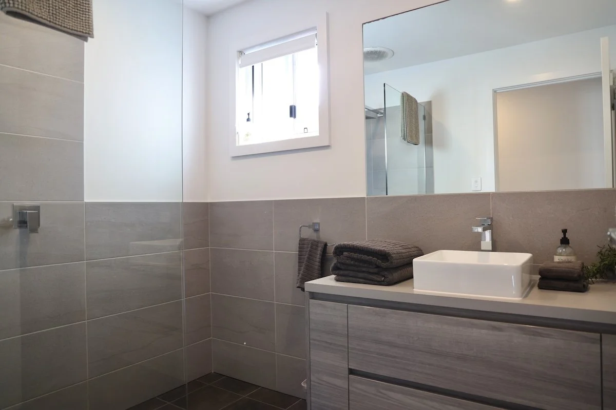 Modern bathroom with a white countertop sink, dark gray towels, a soap dispenser, and a large mirror. A small window provides natural light, and a glass shower enclosure is partially visible.